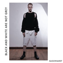 Front view. Man with short hair and beard ist wearing a huge handknitted black pullover with cut outs at the shoulders and long sleeves. He also wears white shorts ad black boots. He stands on a wooden floor and in front of a white wall.