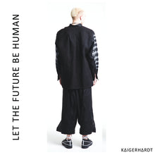 White man with blond hair wears a black shirt with white placed screen prints. The back of the shirt is black without any prints and has  asymetrical fold and seam.  He also wears a black and white pair of 3/4 trousers in hakama style and black shoes with white shoelaces.