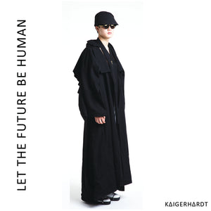 Rigt side view. An asian model wears a heavy, hug black wool coat with an asymetrical collar and a big knitted and felted art piec at the back of the coat. He also wears sunglasses, a black hat, a accessoires around his neck and white shoes with black soles and black shoelaces.
