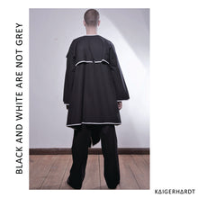 Back view.  A male model with short hair is looking not to the camera. Wearing a big black coat reminding on a draped version of the trench coat with white outlines and black buttons for the kollar. Under the Jacket he wears a black t-shirt, a black pait of trousers and leather shoes. On the right hand the model wears a white enemal ring. He is standing on wooden floor in front of a white wall.