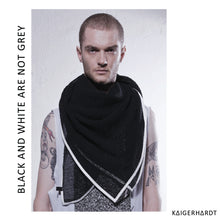 Male model with short hair and beard looking into the camera. Wearing a black scarf out of lightly woven material with white outlines and a white tank top. Both upper arms got tattoos. in the background is a white wall. font left side from down to up: black and white are not grey. font right down corner: kaigerhardt