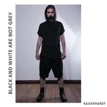 Front view. Man with beard and long hair wears black draped t-shirt with raw edges, black shorts and black leather boots. He is starring into the camera.