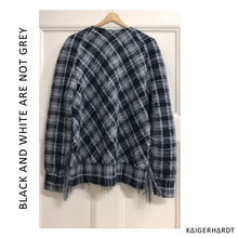 Back view. Checkered black and white sweater withe fringes and raglan sleeves on a hanger hanging on a door. font left side from down to up: black and white are not grey. font right down corner: kaigerhardt