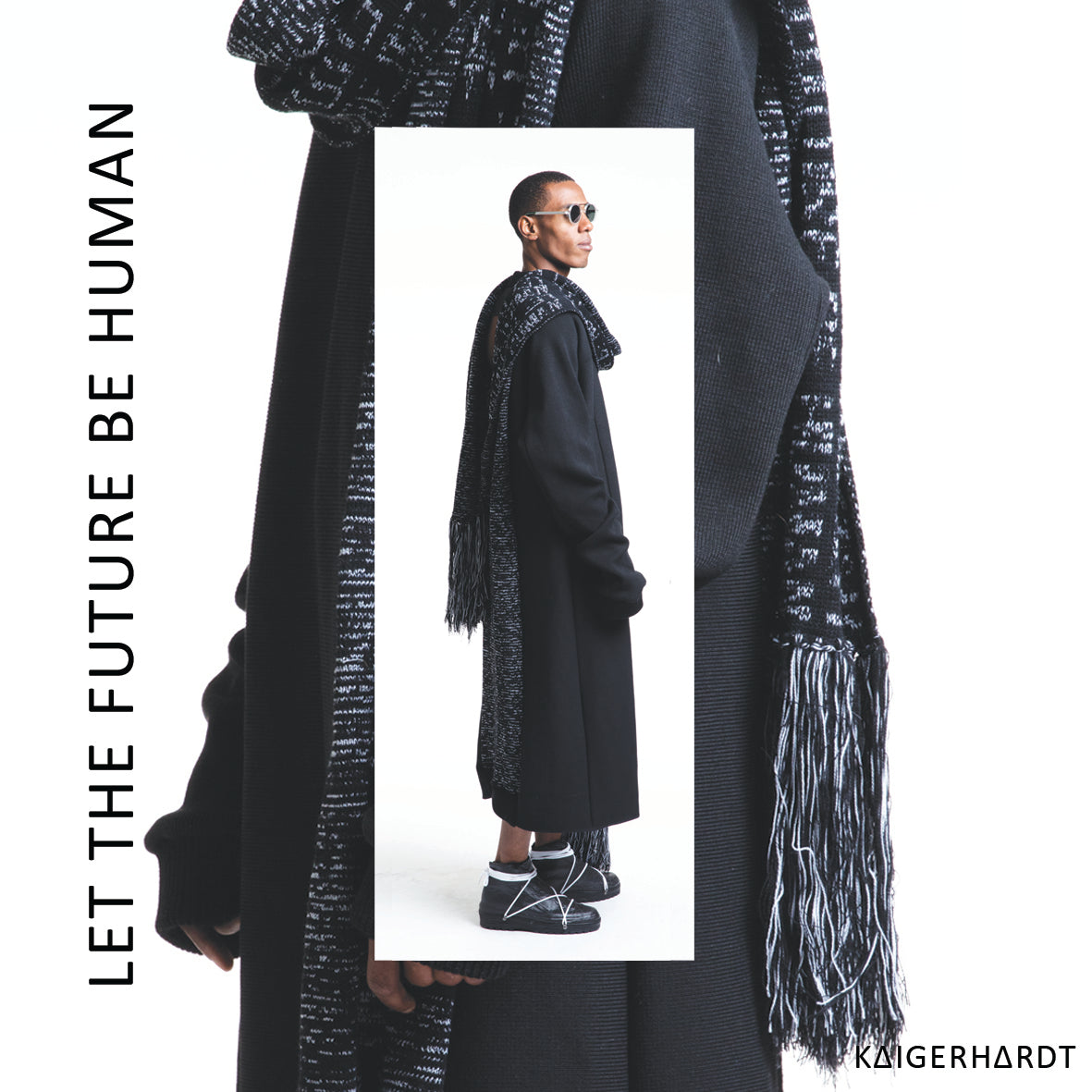 A man wears a black dress in heavy knit with an insert scarf piec in black and whit knit with letters which says LET THE FUTURE BE HUMAN. The man is a people of color and wears also sunglasses and black shoes with white shoelace.