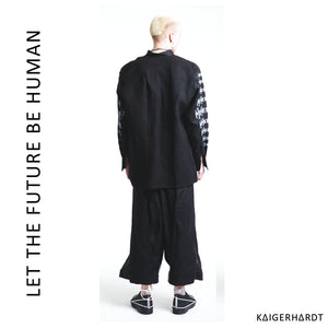 White man with blond hair wears a black shirt with white placed screen prints. The back of the shirt is black without any prints and has  asymetrical fold and seam.  He also wears a black and white pair of 3/4 trousers in hakama style and black shoes with white shoelaces.