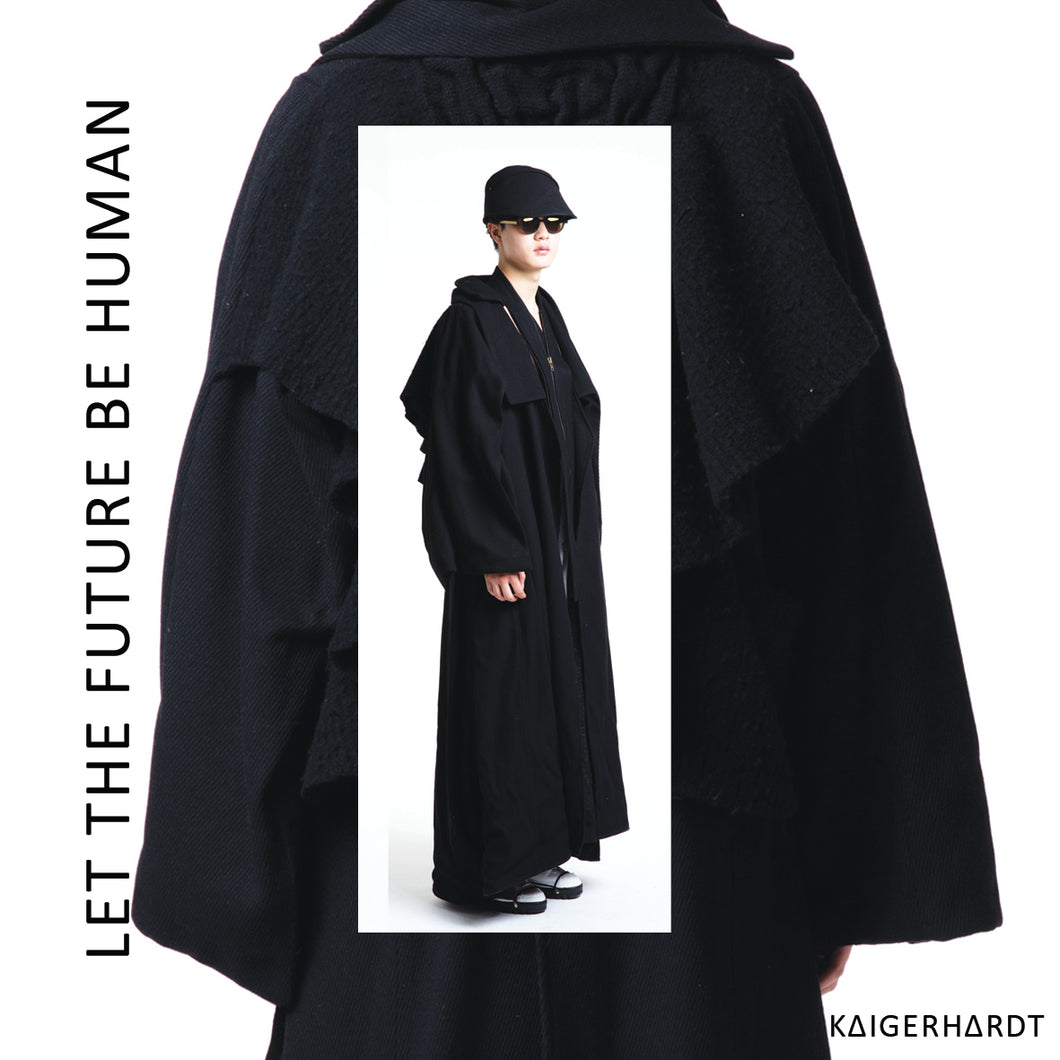 An asian model wears a heavy, hug black wool coat with an asymetrical collar and a big knitted and felted art piec at the back of the coat. He also wears sunglasses, a black hat, a accessoires around his neck and white shoes with black soles and black shoelaces.